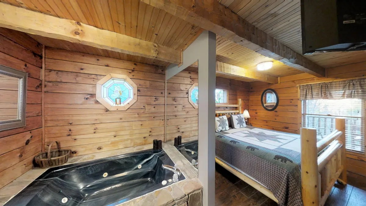 A spacious master suite featuring a cozy bed with decorative pillows, a luxurious jacuzzi tub near octagonal windows, and warm wooden walls. Soft natural light fills the room, creating a restful ambiance. The setting is ideal for unwinding, reading, or enjoying a relaxing soak after a day of adventure.