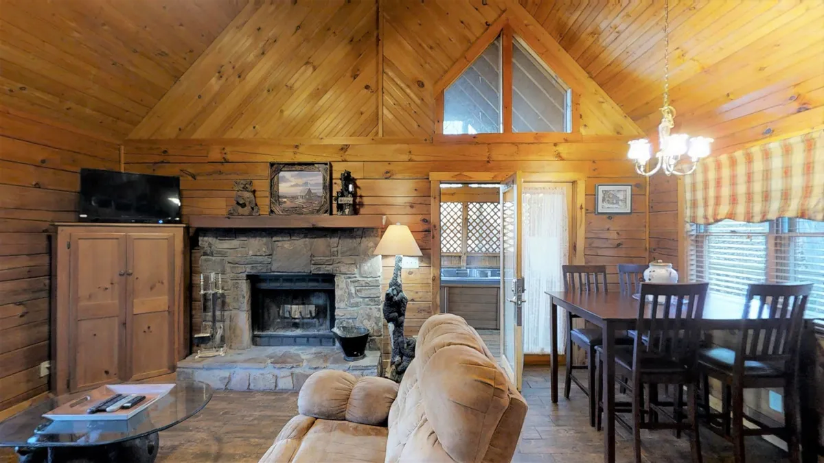 A warm, inviting living room with a prominent wood-burning fireplace, cozy sofa, dining table, and chandelier. Large windows provide natural light and views of the outdoors, while the space is arranged for comfortable gatherings or relaxation. The fireplace takes center stage, emphasizing seasonal fireside comfort, and there's a sense of spaciousness perfect for movie nights or quiet evenings.