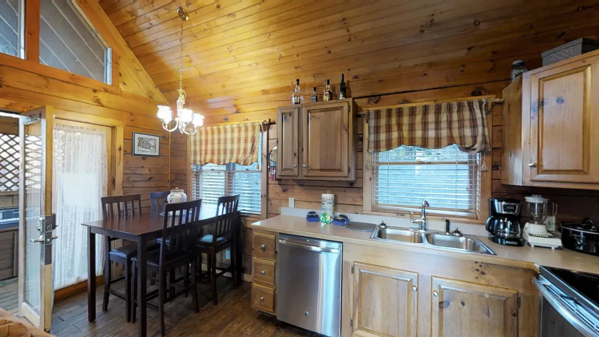 A bright, inviting scene of the kitchen and dining area, with a wooden dining table set for four, a prominent kitchen island, and elegant wooden cabinetry. Sunlight streams in through the windows, highlighting the open layout and sociable atmosphere.