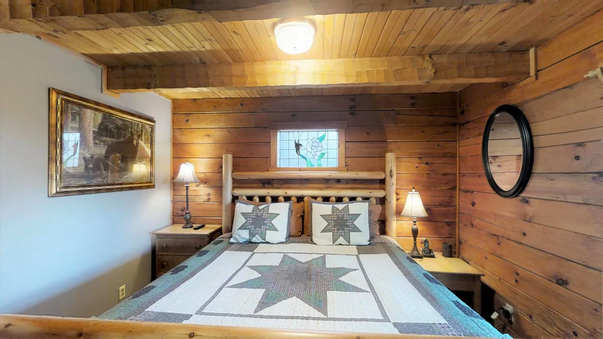 A welcoming bedroom with a log-frame king bed, a colorful quilt, a nightstand topped with a lamp, and a stained glass window letting in gentle morning light. The space feels warm, inviting, and perfect for restful nights or quiet mornings with a view.