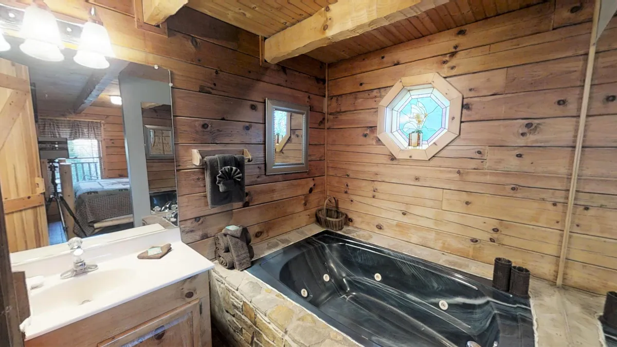 A tranquil bathroom scene with a large jacuzzi tub set beneath a stained glass window, complemented by warm wooden walls, a stylish mirror, and a convenient sink. The interplay of natural light and stained glass adds a serene, spa-like atmosphere ideal for self-care and unwinding.